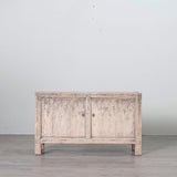 A rustic, light gray distressed wood sideboard cabinet with two doors and small metal knobs, set against a plain light gray wall and gray floor.