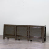 A dark gray wooden sideboard or credenza with three cabinet doors, shown in a minimalist room with a light gray wall and gray floor.