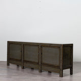 A dark gray wooden sideboard cabinet with three paneled doors, shown in a minimalist room with a light gray wall and gray floor.