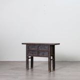A dark brown wooden console table with four drawers and carved drawer fronts, set against a plain light gray wall and gray floor.