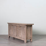 A rustic carved wooden chest or sideboard with a weathered gray-brown finish, set against a plain light gray wall and gray wood floor.