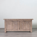A rectangular carved wooden chest or sideboard with three front panels featuring intricate geometric and floral patterns, shown against a plain light gray wall and gray floor.