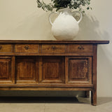 Wooden sideboard with a white vase and greenery against a plain wall.