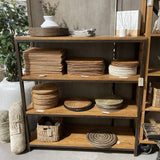 A rustic wooden display shelf unit filled with woven home decor items, including plates, baskets, placemats, and a white ceramic vase, photographed in a store interior.