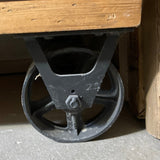 Close-up of a black cast iron industrial wheel under a wooden platform or table on a gray concrete floor.
