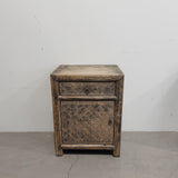 A rustic, vintage-style wooden bedside with a single drawer and a lower cabinet door featuring woven rattan panels, set against a plain white wall and concrete floor.