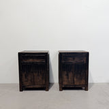 Two dark brown distressed wooden nightstands with one drawer and one double door, set against a plain white wall and concrete floor.