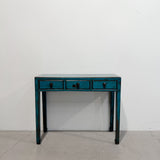A distressed teal blue wooden console table with three drawers and dark metal handles, set against a plain white wall and concrete floor.