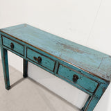 A rustic distressed console table with a weathered turquoise blue painted finish and three drawers, photographed in a simple indoor setting.