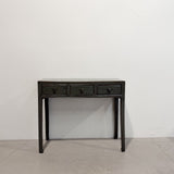 A dark, distressed wooden console table with three drawers and small round knobs, set against a plain white wall and concrete floor.