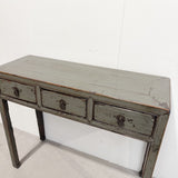 A rustic three-drawer console table with a distressed sage green painted finish and dark metal drawer pulls, shown against a plain white wall.