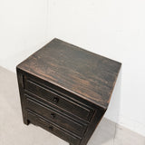 A rustic dark brown wooden nightstand with three drawers and distressed black metal knobs, photographed in a minimalist white room setting.