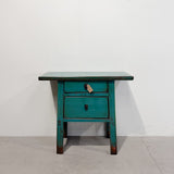 A rustic turquoise painted wooden nightstand with two drawers and a small hanging tag, set against a plain white wall and concrete floor.