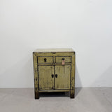 A distressed vintage-style wooden cabinet with a light olive green paint finish and dark weathered detailing, featuring two small top drawers and two lower doors with dark metal knobs, set against a plain white wall and concrete floor.