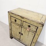 A distressed vintage-style wooden cabinet with a weathered yellow-beige paint finish, black metal drawer pulls, and a small hanging tag, photographed in a warehouse setting.