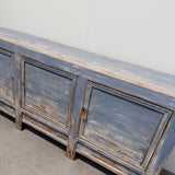 A rustic distressed blue wooden sideboard cabinet with three doors, showing weathered brown wood underneath the faded blue paint, photographed against a plain light gray wall and concrete floor.