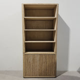A tall, narrow wooden bookcase with four open shelves and a slatted lower cabinet door, shown against a plain light gray wall and floor.