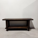 A dark brown wooden console table with a top shelf, photographed against a plain white wall and light concrete floor.