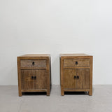 Two matching rustic wooden nightstands with a single drawer and double door, shown side by side against a plain white wall and concrete floor.