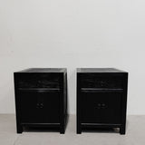 A pair of black wooden nightstands with a single drawer and two cabinet doors, set against a plain white wall and concrete floor.