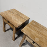 Two rustic light brown wooden side tables with single drawers and black metal ring pulls, shown on a concrete floor against a plain off-white wall.