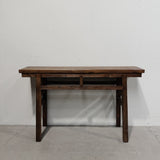 A dark brown wooden console table with a single drawer and open shelf, set against a plain white wall and concrete floor.