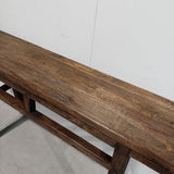 A close-up of a dark brown wooden bench with a glossy finish, showing the natural wood grain and traditional support structure against a plain white wall.
