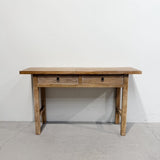 A rustic wooden writing desk with two drawers and small black metal knobs, set against a plain white wall and gray concrete floor.