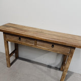 A rustic wooden console table with two drawers and black metal ring pulls, photographed against a plain white wall and concrete floor.
