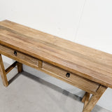 A rustic wooden console table with two drawers, featuring a natural oak-like finish and small black metal knobs, photographed against a plain white wall and concrete floor.