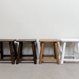 Three wooden stools of varying heights and finishes lined up against a plain off-white wall and concrete floor, with a fourth white stool partially visible on the right.