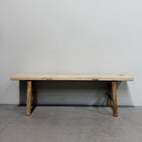 A rustic wooden bench with a light natural wood top and weathered brown legs, positioned against a plain gray wall and concrete floor.