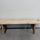 A rustic wooden bench made from light-colored reclaimed wood planks, supported by two angled wooden legs, set against a plain gray concrete floor and white wall.