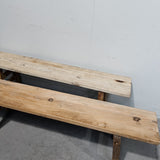 A rustic two-seater wooden bench made from two light natural wood planks on a simple wooden frame, photographed in a warehouse setting against a plain light gray wall.