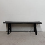 A long black wooden bench with a slatted seat and A-frame legs, photographed against a plain white wall and light gray concrete floor.