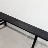 A long black wooden bench with a dark wood-grain finish and black metal legs, positioned against a white wall on a light gray concrete floor.