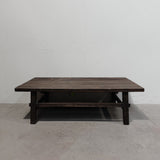 A dark brown wooden coffee table with a rustic finish, featuring a thick rectangular top, four sturdy legs, and a lower support rail, set against a plain white wall and concrete floor.
