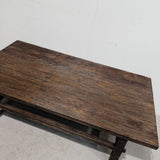 A rustic dark brown wooden coffee table with a thick top and a lower shelf, photographed against a plain off-white wall and light concrete floor.