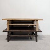 A rustic two-tier wooden coffee table with a light natural wood top and shelves, set against a plain white wall and concrete floor.