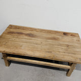 A rustic rectangular wooden coffee table with a natural weathered finish, shown from an elevated angle against a plain off-white wall and concrete floor.