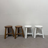 Four stools arranged in a row against a plain white wall and concrete floor, with two wooden stools on the left and two white stools on the right.