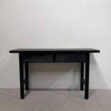 A black wooden console table with two drawers and a rectangular top, positioned against a plain white wall on a concrete floor.