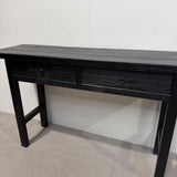 A black wooden console table with two drawers and small round knobs, shown against a plain white wall and concrete floor.