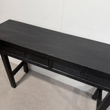 A black wooden console table with two drawers and small round knobs, photographed in a warehouse or showroom setting against a white wall and concrete floor.