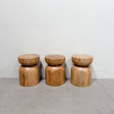 Three rustic wooden stools made from natural wood grain, stacked with a round seat on a cylindrical base, set against a plain white wall and concrete floor.