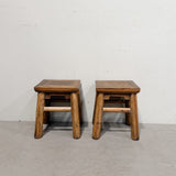 A pair of rustic wooden stools with woven cane seats, positioned side-by-side against a plain off-white wall and concrete floor.