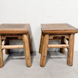 A pair of rustic wooden stools with woven cane seats, shown side by side against a plain white wall and concrete floor.