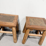 Two rustic wooden stools with woven cane or rattan seats, shown from the side against a plain white wall and gray floor.