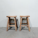 Two rustic wooden stools with a weathered, light brown finish, positioned side-by-side against a plain white wall and concrete floor.