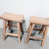 Two rustic wooden stools with curved slatted seats and thick crossed legs, shown on a concrete floor against a plain white wall.
